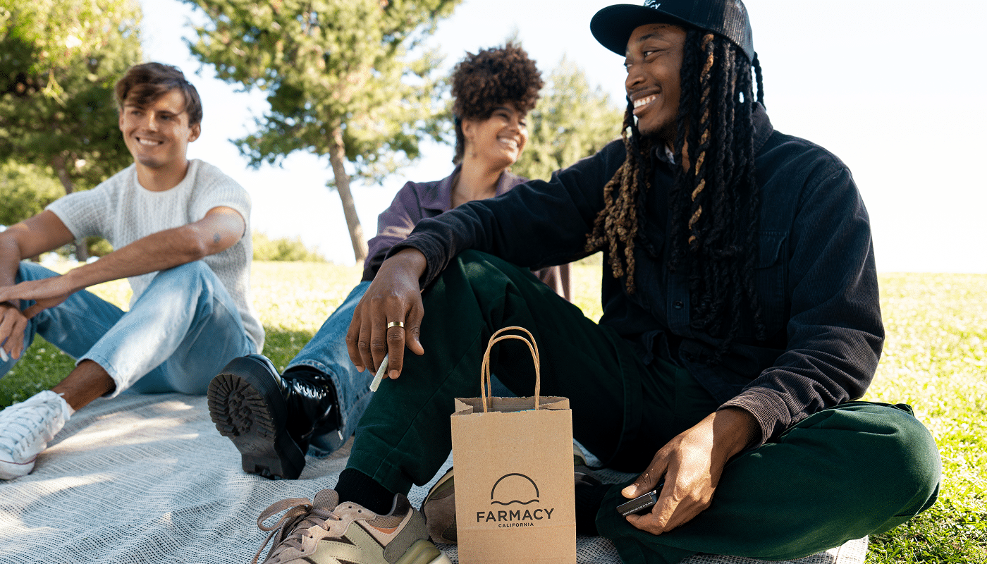 Three friends sitting on a blanket in a grassy park, with a Farmacy California brown paper shopping bag in the foreground.
