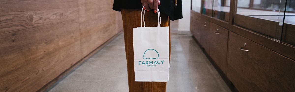 A person holding a white paper shopping bag featuring the Farmacy Berkeley logo in a modern, wood-paneled retail dispensary.