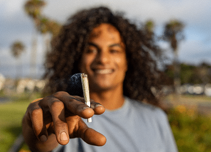 A smiling man with long curly hair holding out a lit pre-roll joint toward the camera in a sunny, outdoor park setting with palm trees.