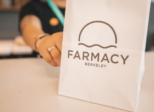 A close-up of a white Farmacy Berkeley shopping bag on a counter, with a staff member's hand reaching out in the blurred background.