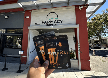 A person holding two black and gold Reeform cannabis product pouches in front of the Farmacy Berkeley storefront entrance.