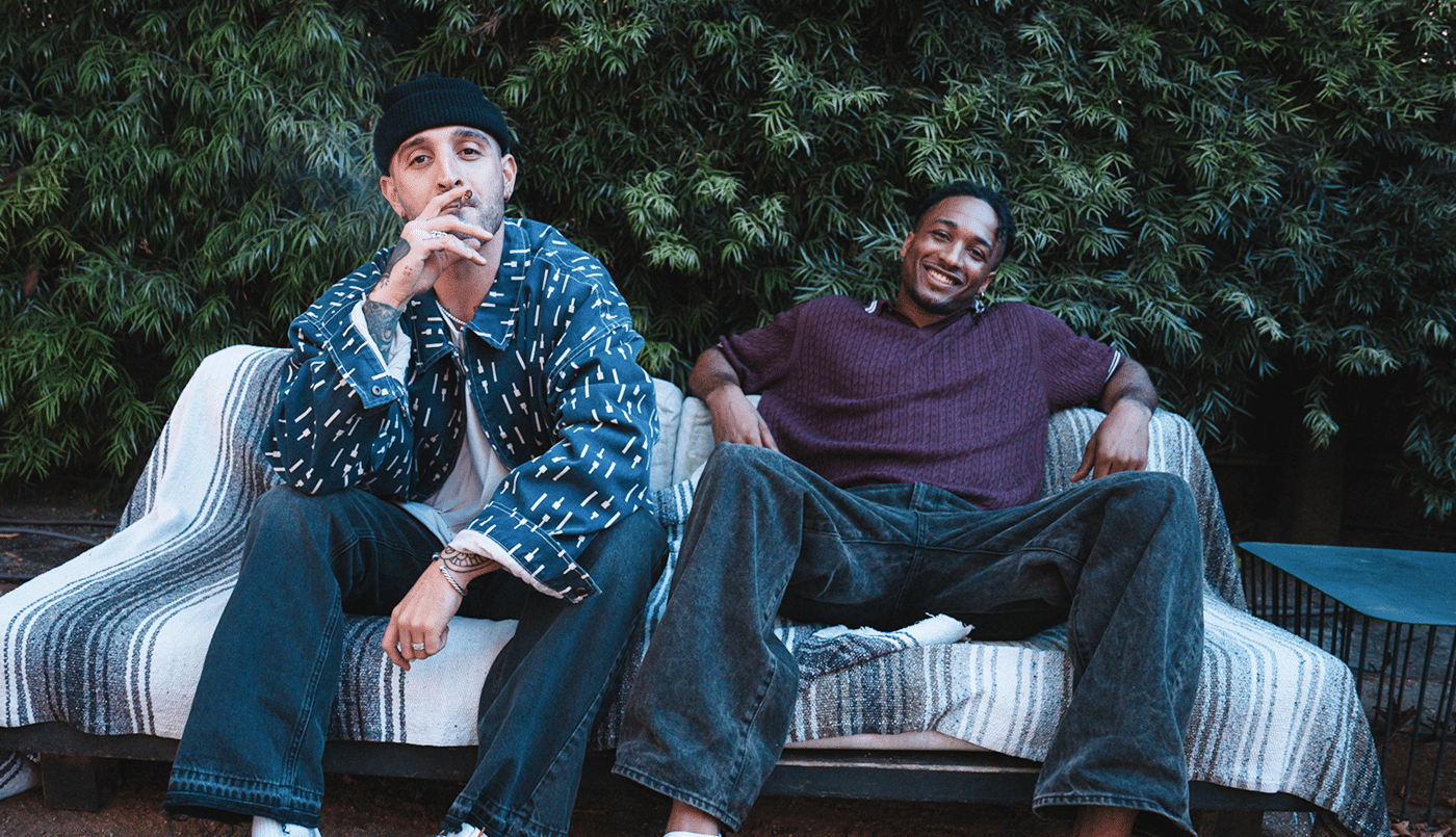 Two men sitting outside on a striped blanket-covered bench, relaxing and smiling in front of lush green foliage.