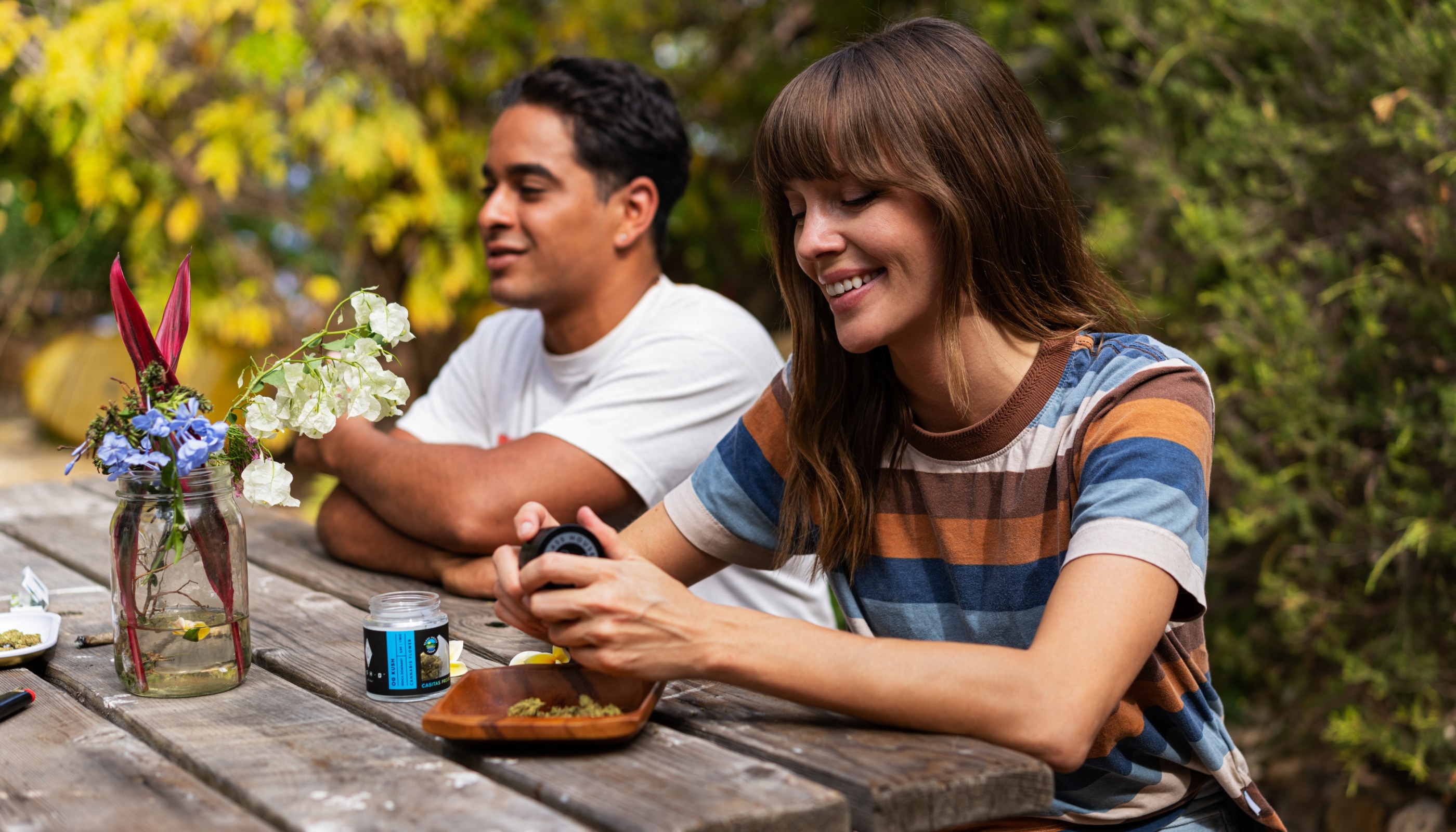 A woman in a striped shirt smiling while preparing cannabis flower at a wooden picnic table outdoors with a friend.