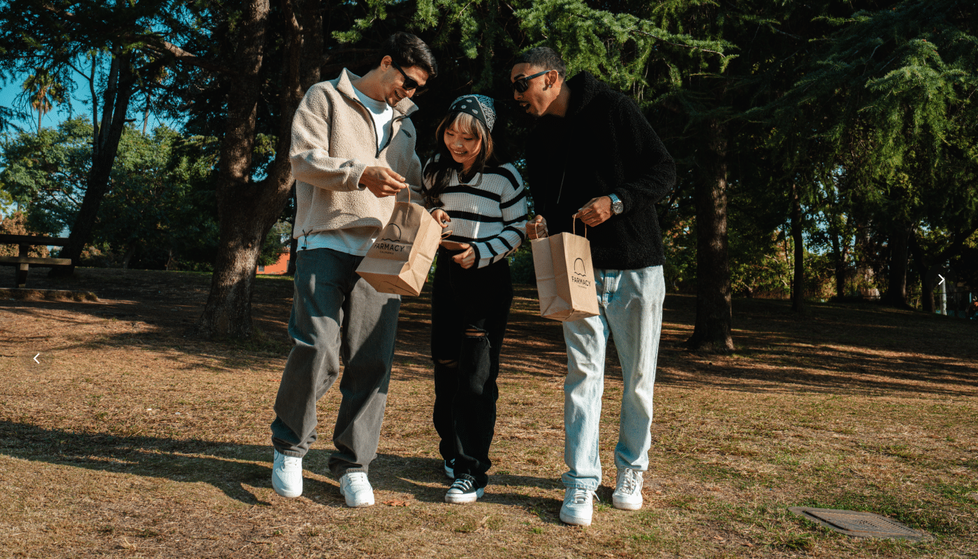 Three friends standing in a sunny, wooded park, smiling and looking into their Farmacy California brown paper shopping bags.