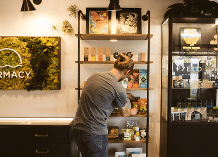 A retail associate in a grey t-shirt organizing high-end cannabis products on modern wooden shelving next to a lush green moss wall featuring the Farmacy logo.