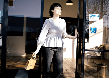 A smiling delivery person walking out of the glass front doors of a Farmacy dispensary while holding a small brown paper shopping bag.